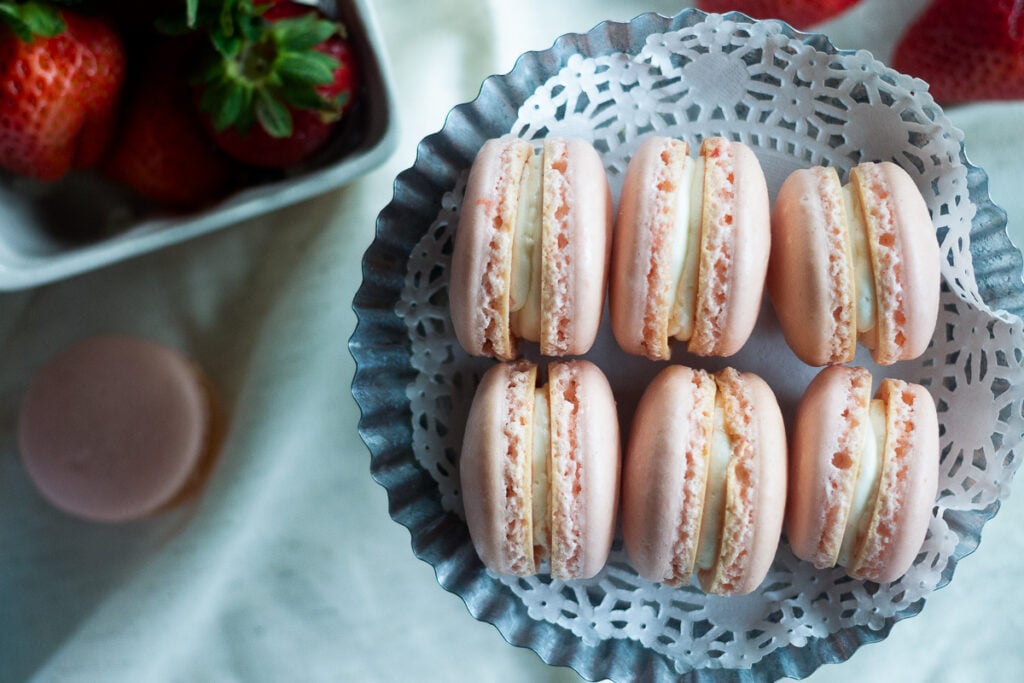 Strawberry macarons with vanilla buttercream filling on mini silver cake plate lined with doily.