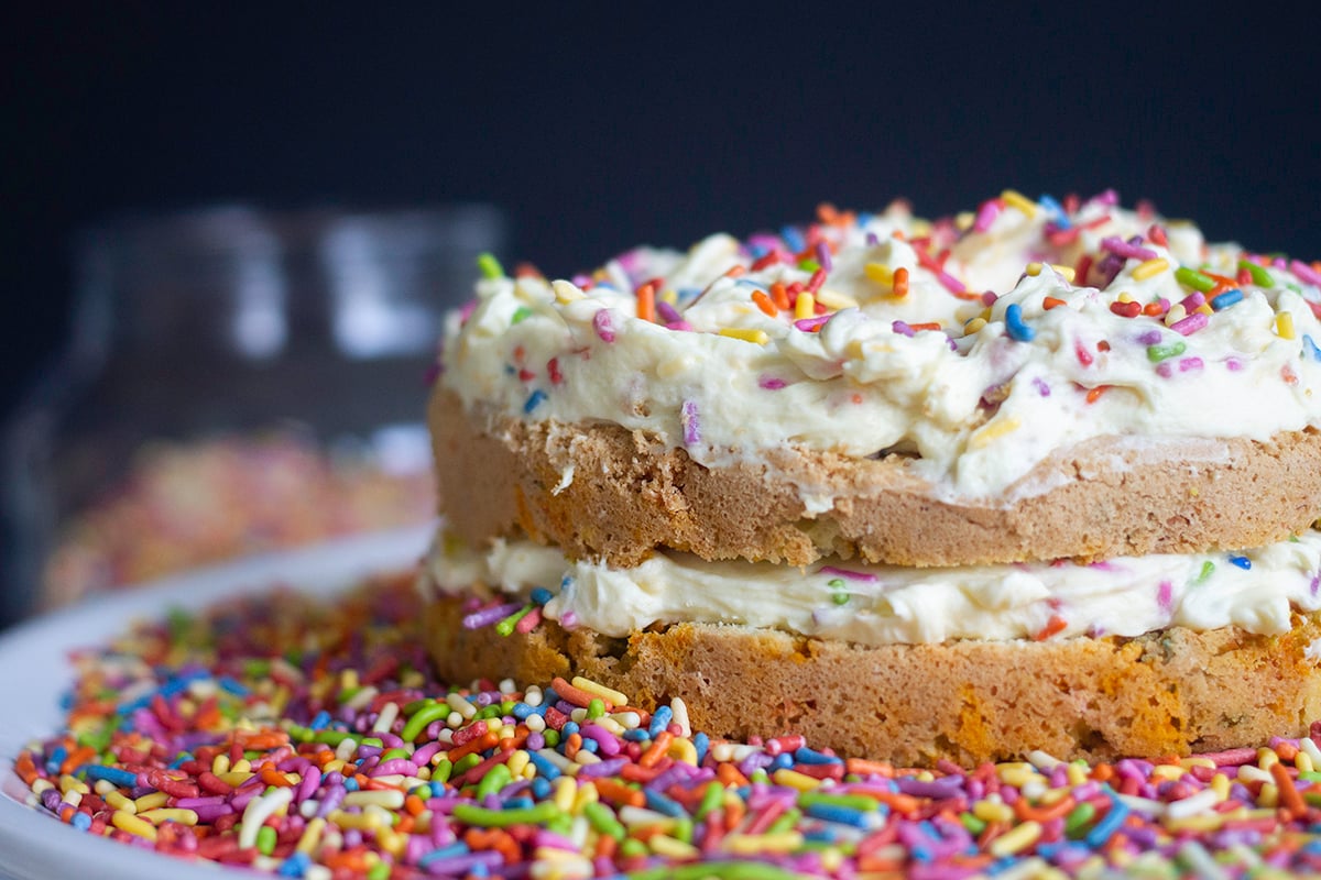 A naked vanilla confetti cake topped and surrounded by rainbow sprinkles.