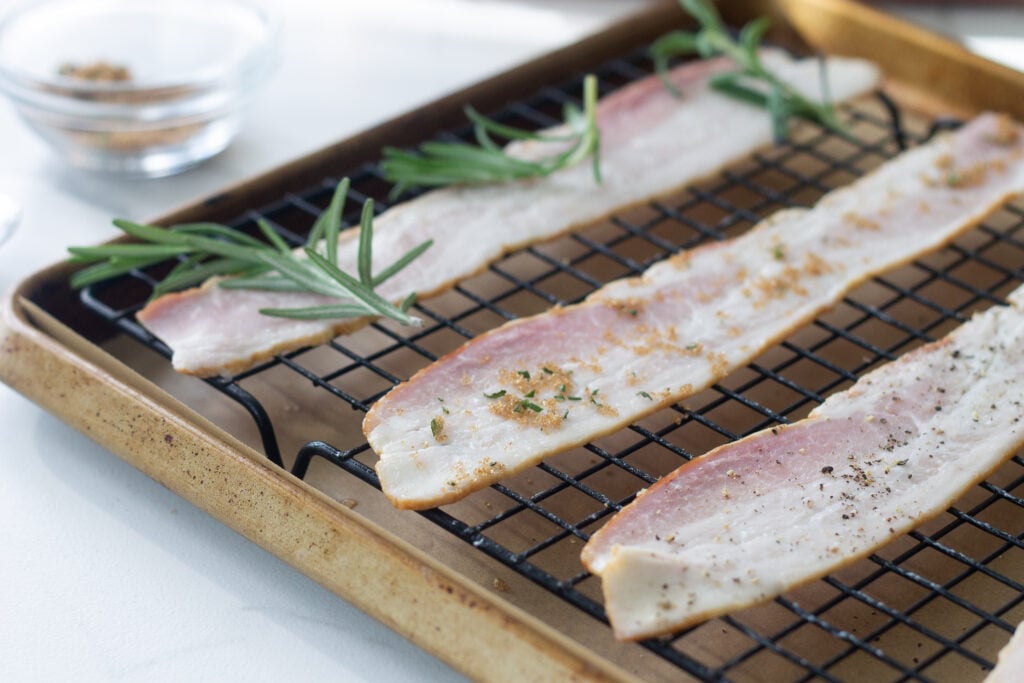 Three bacon slices on sheet pan sprinkled with rosemary, black pepper and sugar and chili powder.