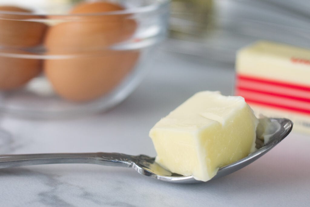 Heaping tablespoon of butter on spoon on counter in front of bowl of raw eggs.