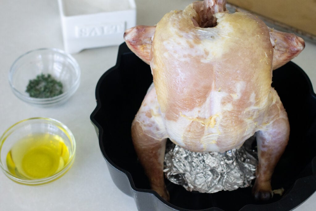 Whole chicken with butt end set over the tube of a bundt pan on countertop next to seasoning ingredients.