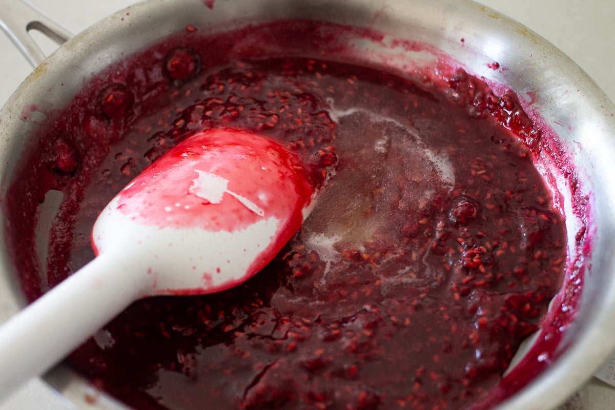 A white spatula stirring raspberry jam in a pan.