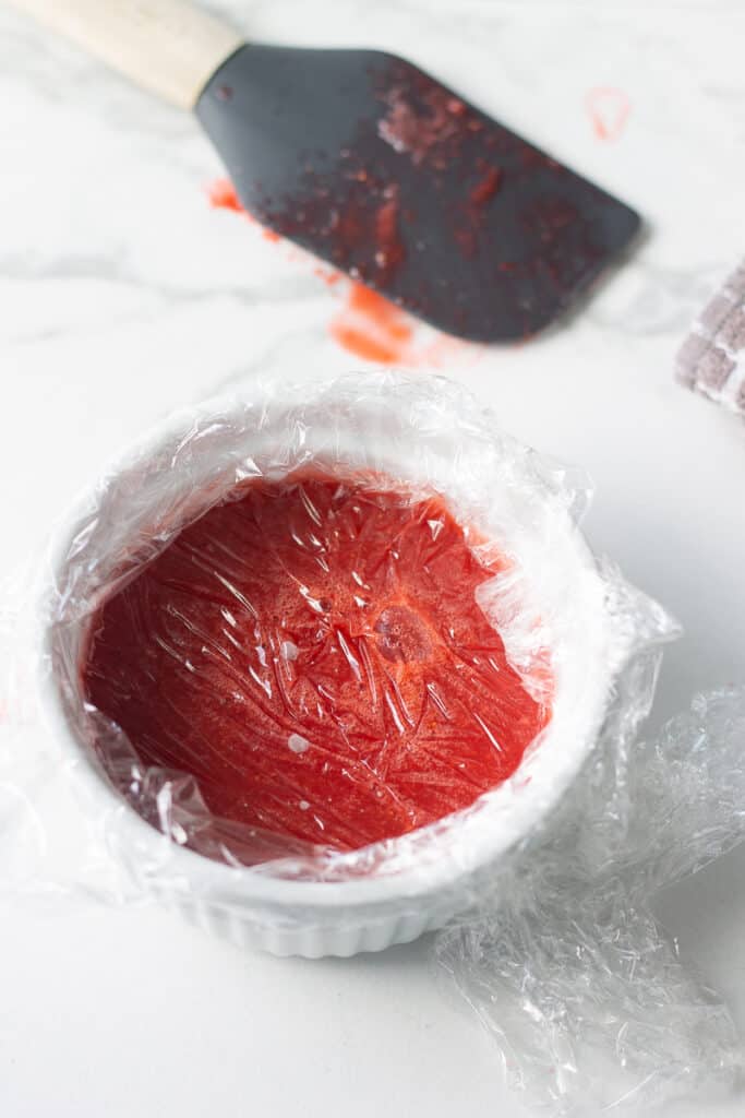 Plastic wrap pressed on top of strawberry jam in a ramekin.