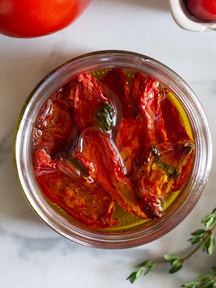 Looking down on a jar of sun-dried tomatoes in oil next to herbs on a counter.