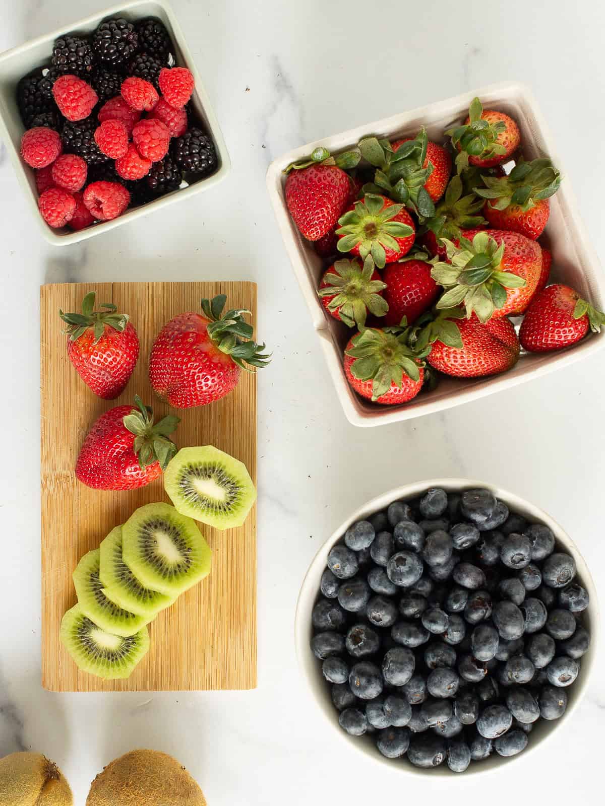 Containers with strawberries, blackberries, blueberries, and a small cutting board with strawberries and kiwi slices.
