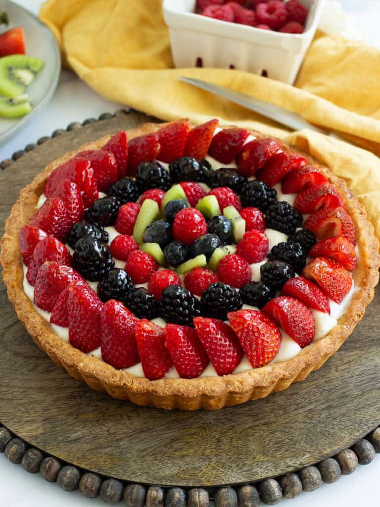 A fresh fruit tart with barries and kiwi on a dark cake plate with a yellow napkin and knife behind.