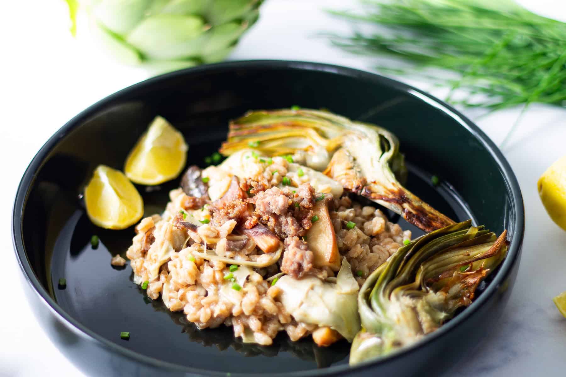 Cooked farro on a black plate with artichokes and lemon wedges.